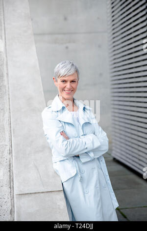 Ritratto di contenuto imprenditrice maturo appoggiata contro la parete indossando azzurro trench Foto Stock