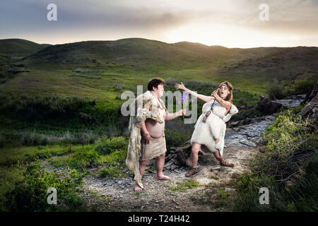 Primitiva caveman vestiti di pelle di animale dando ai fiori di grotta felice donna in montagna Foto Stock