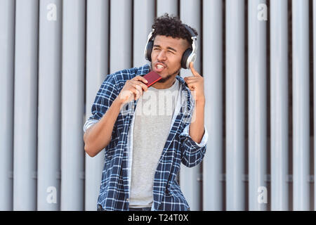Giovane mulatto uomo che indossa le cuffie in piedi isolato su una strada di città ascoltando musica azienda mp3 player cantando cool Foto Stock