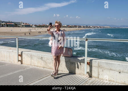 Una donna in un abito beige prendendo un selfie su un molo di cemento con mare e spiaggia Malvarrosa Valencia vista mare Valencia Spagna spiaggia Foto Stock