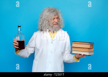 Vecchio con i capelli lunghi uomo che indossa Doctor's vestaglia studio isolato sulla parete blu in piedi tenendo la bottiglia di alcool in una mano guardando alla pila di libri in anot Foto Stock