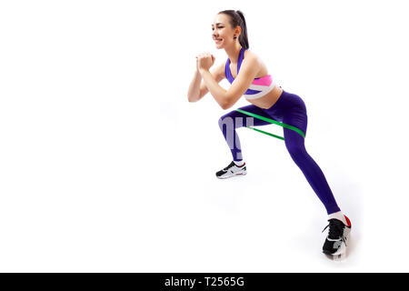 Un dai capelli scuri allenatore donna in sportivo viola corto superiore e palestra mostra la corretta tecnica di squatting con sport fitness bande di gomma; la posizione di Foto Stock