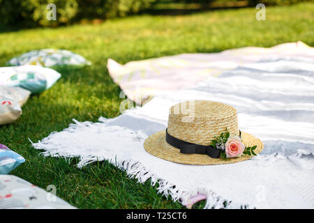 Cappello di paglia con fiori freschi giaceva su un bianco Coperta picnic a prato verde luminoso giorno di estate sfondo. Summer Weekend di piacere. Foto Stock