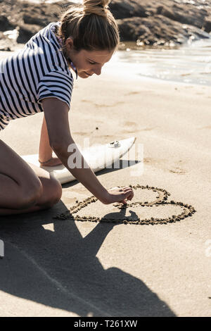 Giovane donna con la tavola da surf di disegno di un cuore nella sabbia Foto Stock