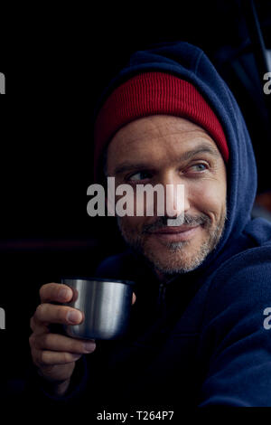 Ritratto di barbuto uomo maturo con la tazza di termos indossa cappuccio rosso e blu giacca con cappuccio Foto Stock