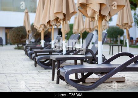 Un certo numero di vuoto spiaggia sdraio vicino alla piscina chiusa sotto gli ombrelloni. Spiaggia di saloni, lasciando in prospettiva. Sedie a sdraio, pronto a prendere un periodo di riposo Foto Stock