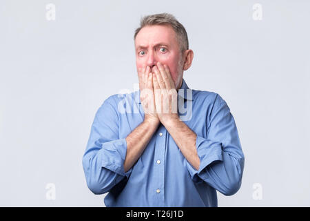 Scioccato senior Uomo in camicia blu copre la bocca e cerca disperatamente verso il basso, avvisi somehing orribile, isolato su sfondo bianco. Studio shoot Foto Stock