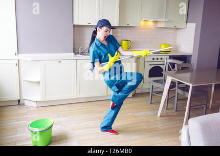 Funny lavoratore dancing in cucina. Lei finge di suonare la chitarra. Ma la ragazza ha mop in mani in stand di che. Ella è oscillante e rilassante Foto Stock