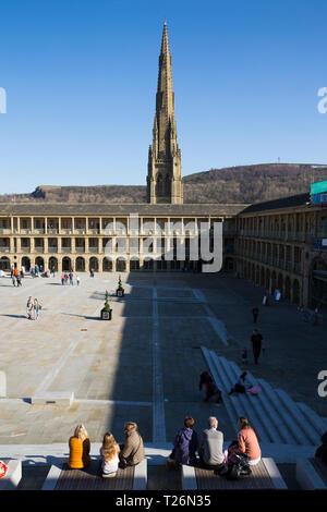 Ai visitatori di rilassarsi sui sedili sedile unico / panche in pezza Hall con piazza Chiesa Cappella guglia dietro in background. Sunny / sun & blue sky. Halifax, West Yorkshire, Regno Unito. (106) Foto Stock