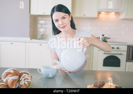 Positivo giovane donna si siede a tavola in cucina e si affaccia sulla fotocamera. Essa detiene il bollitore bianco e versa acqua nella tazza. La donna pone. Lei sembra bello Foto Stock