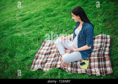 Ritratto di un felice capelli neri e orgogliosa donna incinta nel parco. Il modello femminile è seduto sull'erba ed è scrivere note Foto Stock
