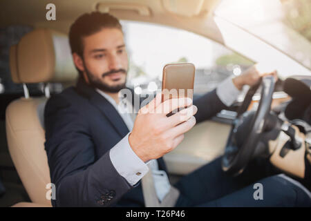 Gentile e simpatico imprenditore si siede in auto di lusso. Egli detiene il telefono in una mano e guarda il suo schermo. Un altro è la mano sul volante Foto Stock