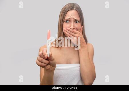 Spaventato e preoccupato per il giovane donna si alza e guarda al test di gravidanza. Essa tiene in mano. Modello coveres la bocca con la mano. Il suo corpo coperto di white t Foto Stock