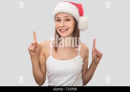 Giovane e donna positivo nel cappello di Natale stand ed i punti verso l'alto. Indossa la t-shirt. Il modello pone. Isolato su sfondo grigio Foto Stock