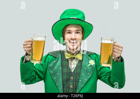 Positivo felice giovane uomo in verde saint patrick suit tenendo due grandi boccali di birra e sorriso. Isolato su sfondo grigio Foto Stock