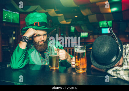 Bevuto giovane uomo in tuta verde sedersi al bancone bar in pub con un amico. Un altro ragazzo si è addormentato. Essi hanno boccali di birra Foto Stock