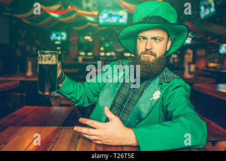 Bevuto giovane uomo in verde San Patrizio la tuta di sedersi a tavola in pub. Egli tenere la tazza di birra scura. Guy refrigerazione Foto Stock