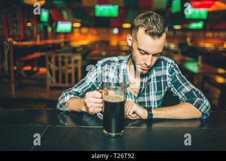 Giovane uomo bello seduto da solo al bancone bar in pub. Egli tenere la tazza di birra e guardare orologi Foto Stock