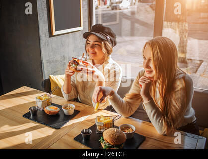 Bella giovani donne siedono in cafe a tavola. Firls ha burger in mani. In attesa di un altro pezzo di patate fritte. Essi godono di mangiare pasti Foto Stock