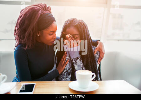 Sconvolto youn donna africana sedersi e grido. Essa copre il viso con le mani. Il suo amico comfort giovane donna. Essi siedono in cafe a tavola Foto Stock