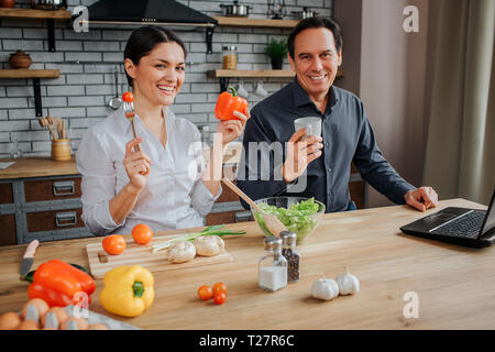 Allegro l uomo e la donna sedersi in cucina e guardare dritto sulla fotocamera. Hanno sorriso. Donna tenere il pomodoro sulla forcella e il pepe in un altra mano. L'uomo ha cup. Essi Foto Stock