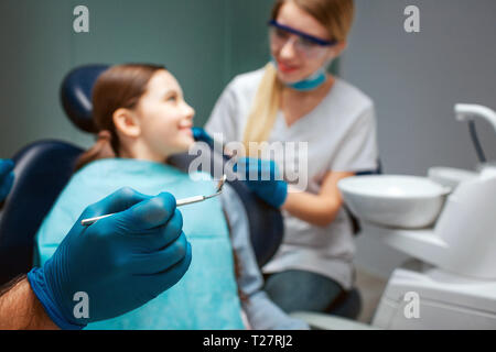 Mano nel guanto in lattice tenere lo strumento dentale tenere lo strumento dentale. Bambino seduto in poltrona e sorriso di donna. Dentista femmina stare accanto a. Foto Stock