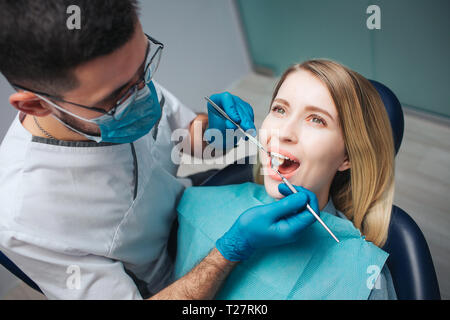 Grave dentista in maschera e tunica bianca accanto a client e controllare la condizioni dei denti. Giovane donna seduti in poltrona e tieni la bocca aperta. Lei guarda in alto. Foto Stock