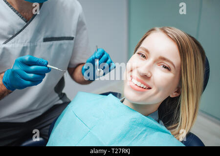 Positivo giovane donna sit in chairin odontoiatria e sorriso alla telecamera. Ella mostra uno splendido sorriso. Medico tenere gli strumenti per il trattamento dei denti. Foto Stock