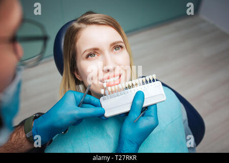 Medico maschio stand su client e mettere i denti artificiali di quelli veri. Procedura per lo sbiancamento dei denti o la sostituzione. Client femmina seduto in poltrona e guardare in alto Foto Stock