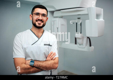 Nizza allegro dentista stand in x-ray camera e pongono sulla fotocamera. Egli ha sorriso. Macchina a raggi X dietro. Foto Stock