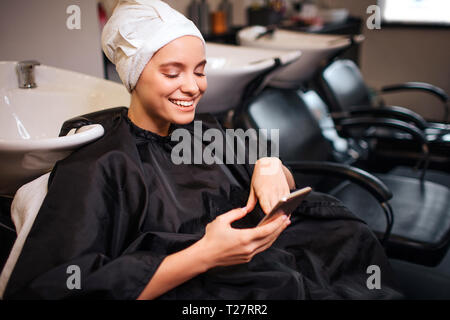 Sorridente bella donna con asciugamano sulla testa guardando il telefono prima di fare l'acconciatura. Hair Spa nel salone di bellezza Foto Stock