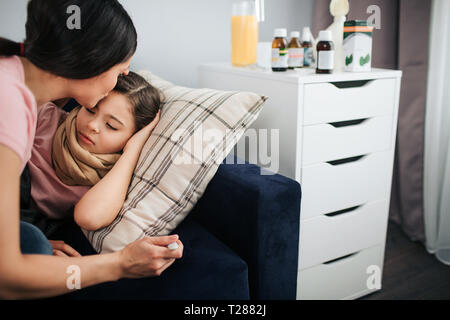 Vista di taglio del giovane donna baciando la fronte di sua figlia. Ella sedersi oltre il suo lettino. Malati ragazza distesa vi. Madre termometro a tenere in mano. Essi sono Foto Stock