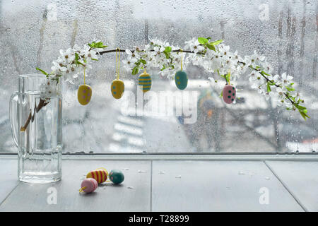 Tavola primaverile con uova di Pasqua sospesi dal rametto coperto con fiori di colore bianco dalla finestra in un giorno di pioggia Foto Stock