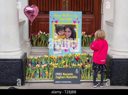 Una giovane ragazza guarda a fiori accanto a un gigante di Giorno della Madre carta lasciata sui passi dell'Ambasciata Iraniana a Knightsbridge, Londra, per carità britannica lavoratore Zaghari-Ratcliffe Nazanin che si terrà in Iran dove lei è accusato di spionaggio. Foto Stock