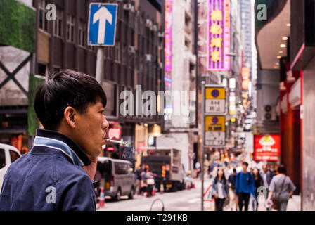 L'uomo, il fumo di sigarette, camminando per strada trafficata, utilizza lo smartphone, close up, Kowloon, Hong Kong Foto Stock