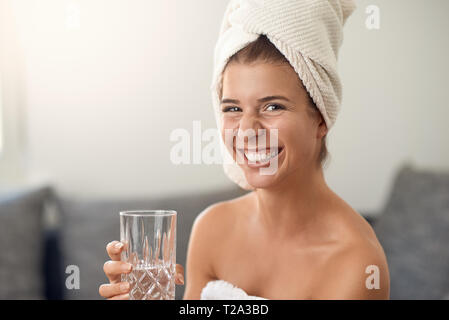 Felice attraente giovane donna con un bel sorriso amichevole indossando un pulito asciugamano bianco intorno i suoi capelli tenendo un bicchiere di pura acqua fresca a ridere a camma Foto Stock