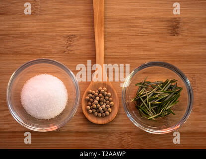 Ciotola di cristallo con il comune sale marino, un cucchiaio di legno con pepe bianco e rosmarino su un tavolo di legno Foto Stock