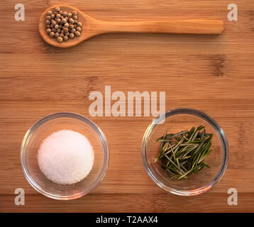 Ciotola di cristallo con il comune sale marino, un cucchiaio di legno con pepe bianco e rosmarino su un tavolo di legno Foto Stock
