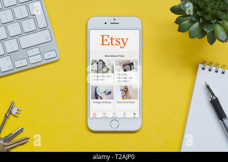 Un iPhone che mostra il sito web Etsy poggia su uno sfondo giallo tabella con una tastiera, tasti, blocco note e di impianto (solo uso editoriale). Foto Stock