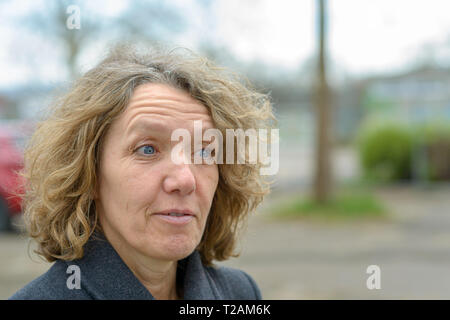 Sorpreso di mezza età con la signora bionda capelli ricci e faccia stropicciata visualizzati nel busto di close-up di mezzo giro ritratto in esterno con spazio di copia Foto Stock