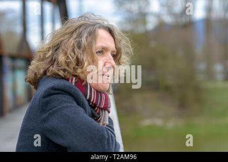 Donna di mezza età con ricci capelli biondi, indossando il cappotto grigio e sciarpa rossa, in piedi sul ponte, appoggiata sulla ringhiera e guardando lontano. lato verticale Foto Stock