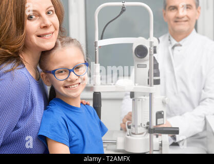 Bella ragazza in blu bicchieri in posa medical oftalmologia clinica con la madre. Coppia medico oculista sorridente, guardando la telecamera, consulenza pa Foto Stock