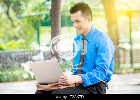 Sorridente imprenditore asiatico seduto e lavorare con il computer portatile con schermo virtuale che visualizzare l'interfaccia del grafico finanziario e mappe del mondo. Digital t Foto Stock