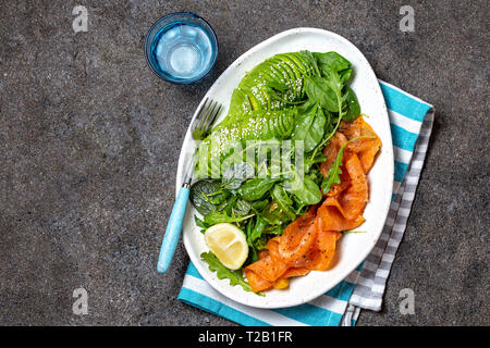 Bassa di un glucide insalata. Gli spinaci, rucola con insalata di avocado e salmone. Nero lo sfondo di calcestruzzo, piastra bianca, vista dall'alto Foto Stock