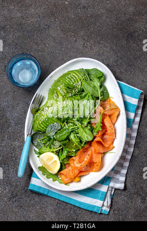 Bassa di un glucide insalata. Gli spinaci, rucola con insalata di avocado e salmone. Nero lo sfondo di calcestruzzo, piastra bianca, vista dall'alto Foto Stock