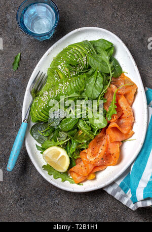 Bassa di un glucide insalata. Gli spinaci, rucola con insalata di avocado e salmone. Nero lo sfondo di calcestruzzo, piastra bianca, vista dall'alto Foto Stock