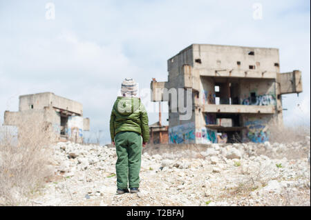 Un piccolo bambino in giacca verde in piedi su ruderi di edifici distrutti in zona di guerra Foto Stock