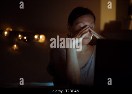Stanco della donna lavoro straordinario sul computer portatile durante la notte nel suo ufficio a casa, viso è illuminato dalla luce blu dello schermo Foto Stock