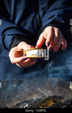 Una ragazza si prepara un sandwich da cookie con marshmallows riscaldato su un incendio nel grill. Dessert picnic Foto Stock