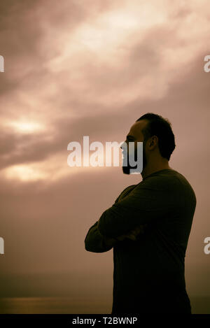 Silhouette di un uomo barbuto in piedi sulla spiaggia al tramonto le braccia incrociate che guarda lontano Foto Stock